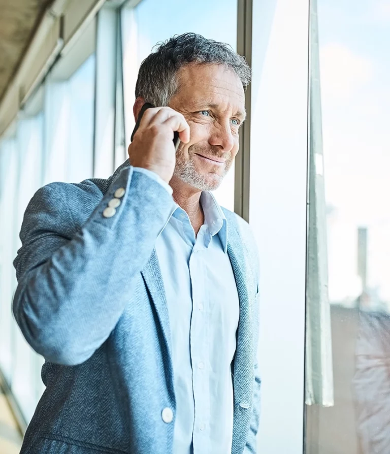 homme mature cheveux gris, regard serein, au téléphone devant une fenêtre d'un édifice à bureau